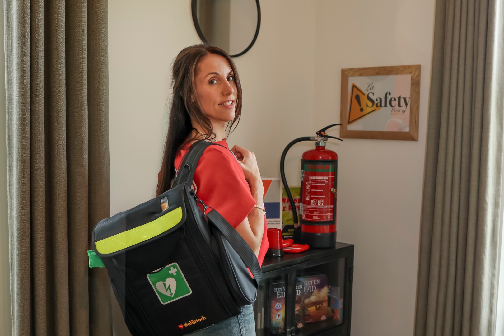 Een glimlachende vrouw van Learncall 112 draagt een zwarte tas met AED-logo in een ruimte met een brandblusser en 'Safety First' bordje.